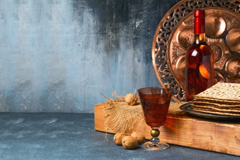 Ein Glas Rotwein, eine Flasche, Walnüsse, Matze und ein dekorativer Seder-Teller sind auf einem Holzbrett vor einem strukturierten blau-grauen Hintergrund angeordnet.